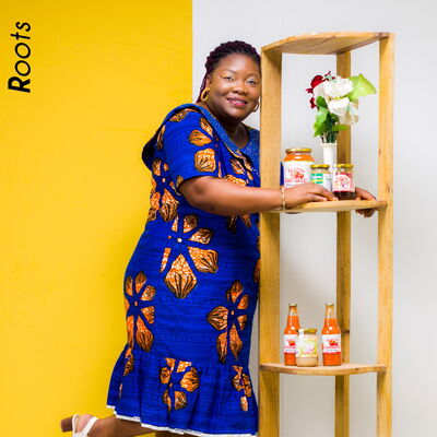 Eine Frau in buntem Kleid posiert lächelnd neben einem Regal mit Gläsern und Blumen vor einer gelben Wand.