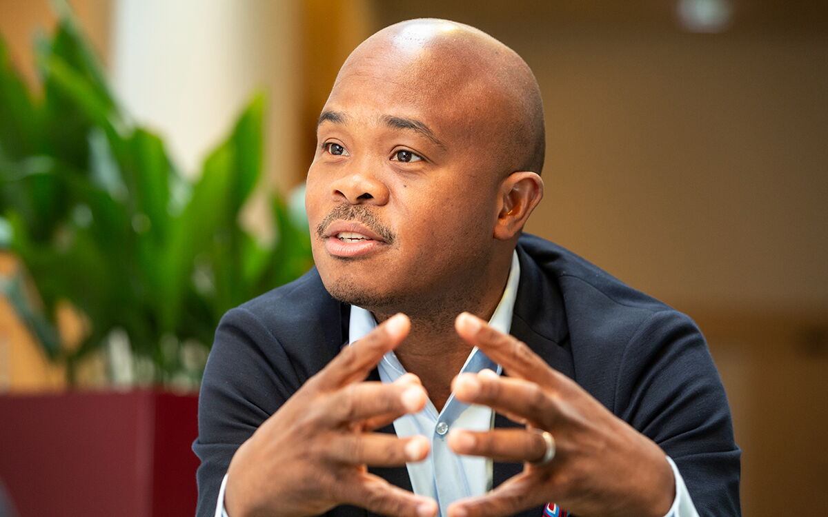 Ein Mann in einem Anzug spricht gestikulierend, während Pflanzen im Hintergrund zu sehen sind.