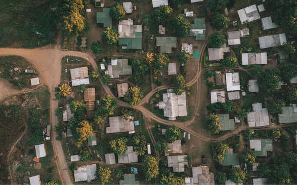 Luftaufnahme eines Dorfes mit dicht beieinander liegenden Häusern und grünen Bäumen entlang der staubigen Straßen.