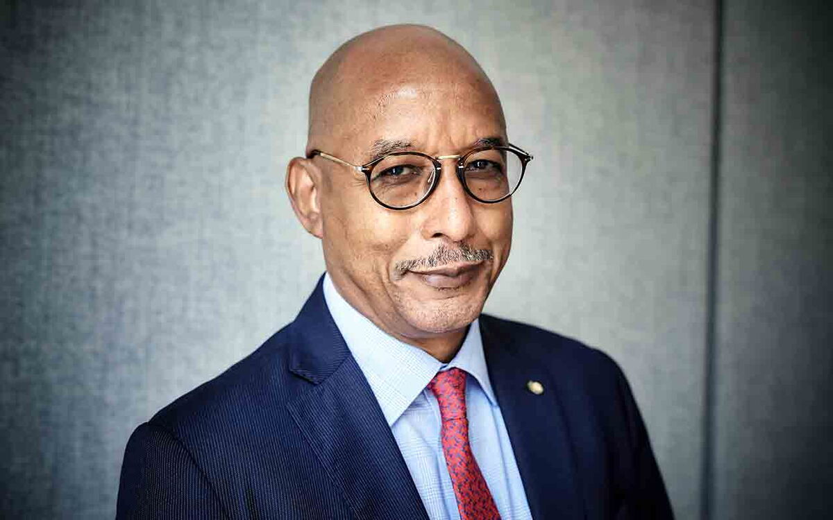 Ein Mann in Anzug und roter Krawatte lächelt freundlich, während er zur Kamera blickt.