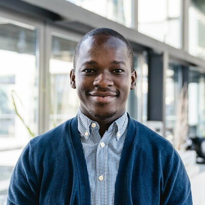 Ein lächelnder Mann in einem blauen Pullover steht in einem modernen Bürogebäude.