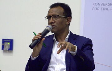 A man in glasses and a suit is speaking into a microphone, gesturing with his hand during a discussion.
