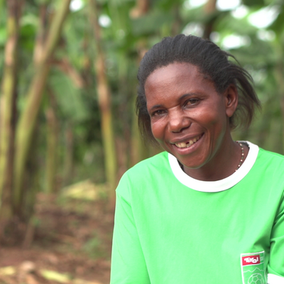 A woman in a green shirt smiles warmly while standing in a lush garden, surrounded by tall green plants.
