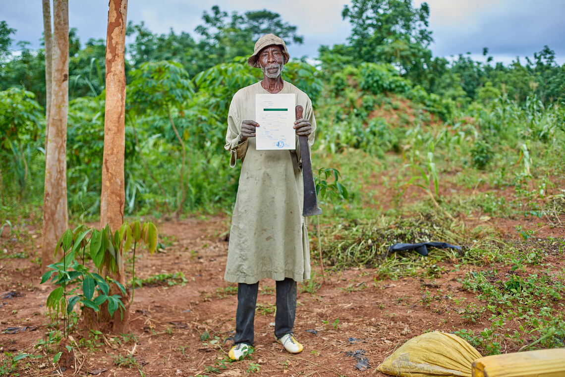 A man stands proudly on his farm, holding up a land certificate amid lush greenery.