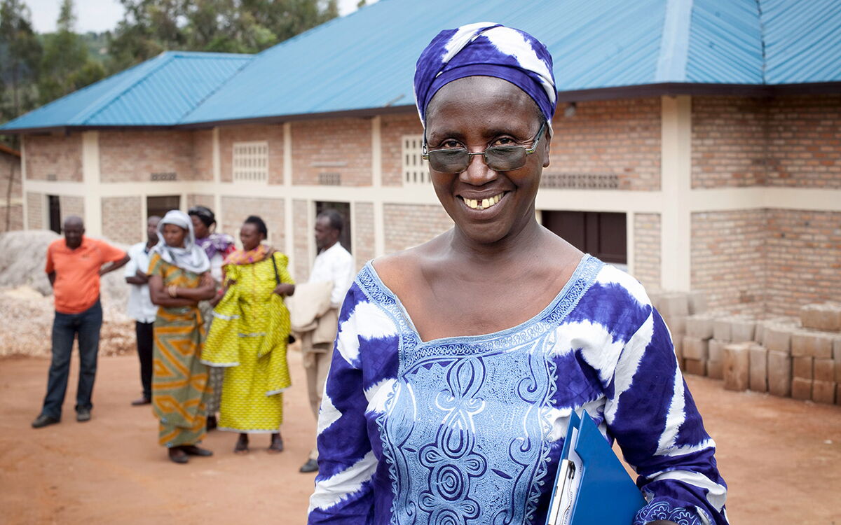 A smiling woman in a vibrant blue patterned outfit stands confidently holding documents, with a group gathered in the background.