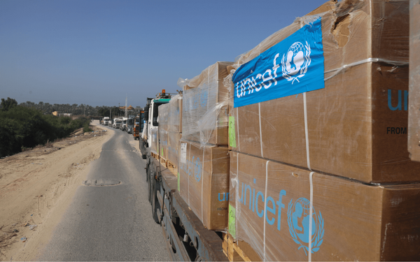 Ein Konvoi von Lastwagen transportiert UNICEF-Hilfsgüter auf einer Landstraße bei sonnigem Wetter.