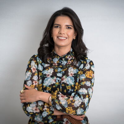 Eine Frau in einem geblümten Kleid steht lächelnd mit verschränkten Armen vor einem neutralen Hintergrund.
