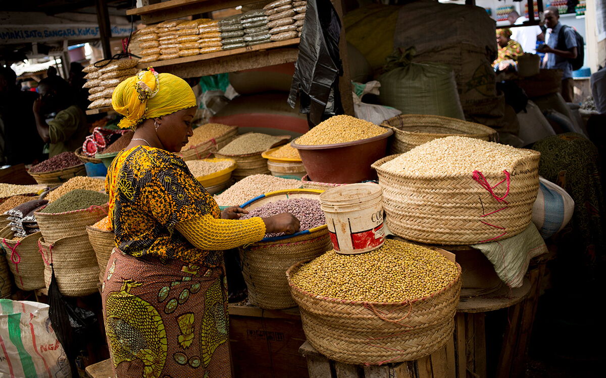 Eine Frau in bunter Kleidung betrachtet Körbe voller Getreide auf einem geschäftigen Markt.