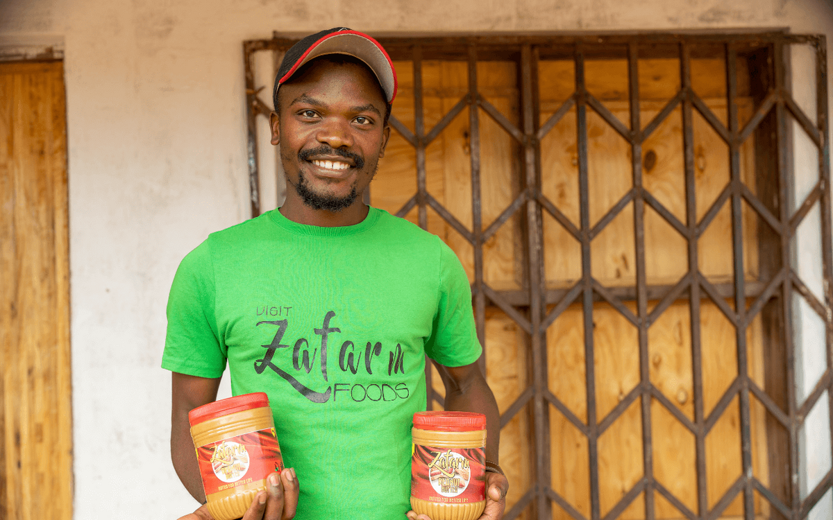 Ein junger Mann in grünem T-Shirt hält zwei Lebensmittelbehälter stolz vor einem Holztor.