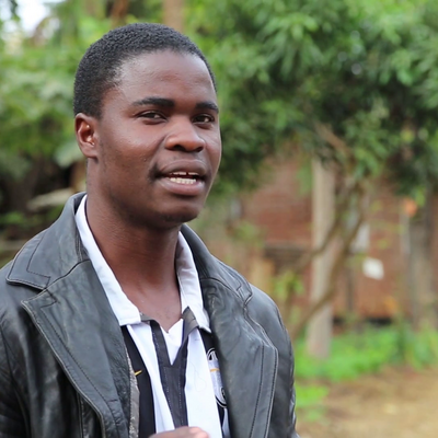 Ein Mann in einer schwarzen Lederjacke spricht draußen vor einem Hintergrund mit Bäumen und unscharfem Gebäude.