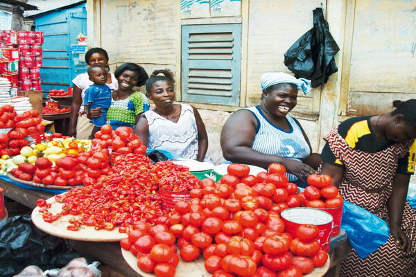Women in a vibrant Ghanaian market smile while selling fresh tomatoes and peppers at their colorful stall.