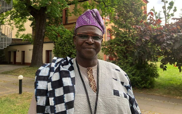 Ein Mann in traditioneller Kleidung und lila Hut steht lächelnd in einem grünen Garten vor einem Backsteingebäude.