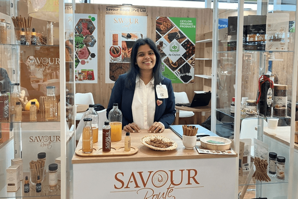 Eine Frau steht lächelnd an einem Messestand von Savour Route mit Getränken und Gewürzen.