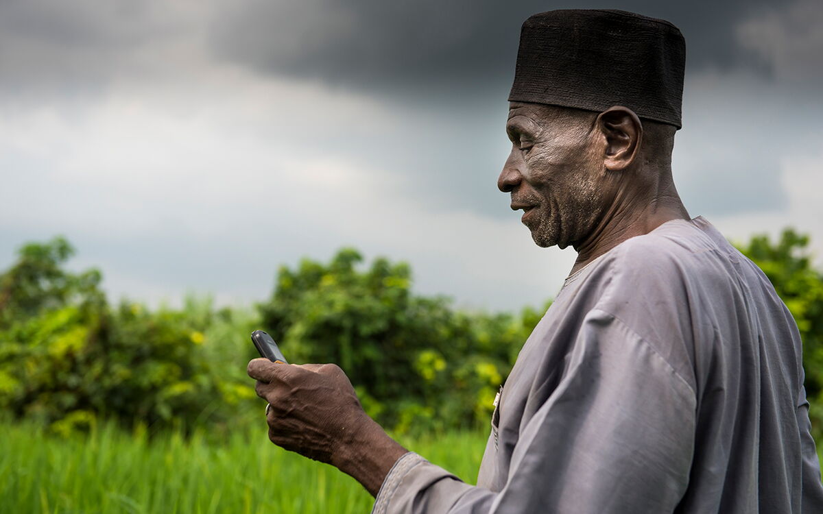 An elderly man in traditional attire uses a mobile phone against a backdrop of lush greenery and cloudy skies in Nigeria.