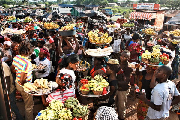 Vibrant scene in Abidjan market with vendors balancing colorful fruits and vegetables on trays amid bustling crowds.