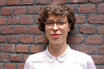 A person with curly hair and glasses stands against a brick wall, wearing a white shirt.