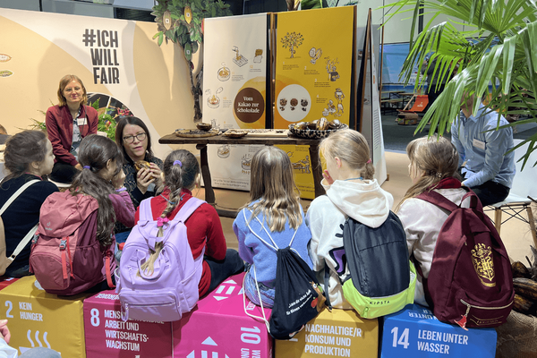 A group of children with backpacks attentively listen to a woman explaining the chocolate-making process at an educational fair.