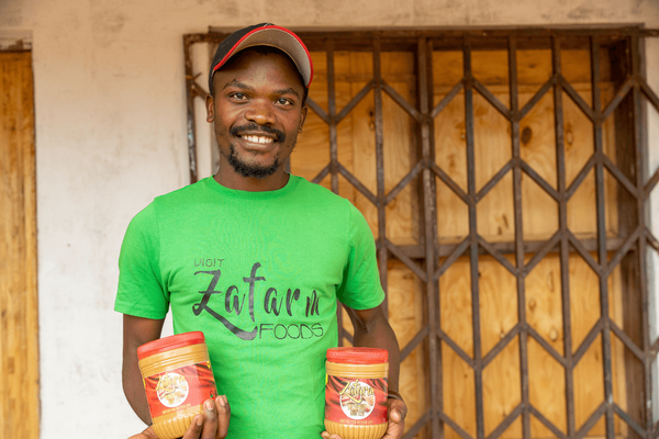 A smiling young man in a green shirt holds two jars of food products in front of a wooden and metal gate.