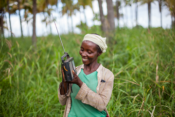 Eine Frau in Tanzania lächelt, während sie ein Radio in einem grünen Feld hält. | © Frederic Courbet für Farm Radio International