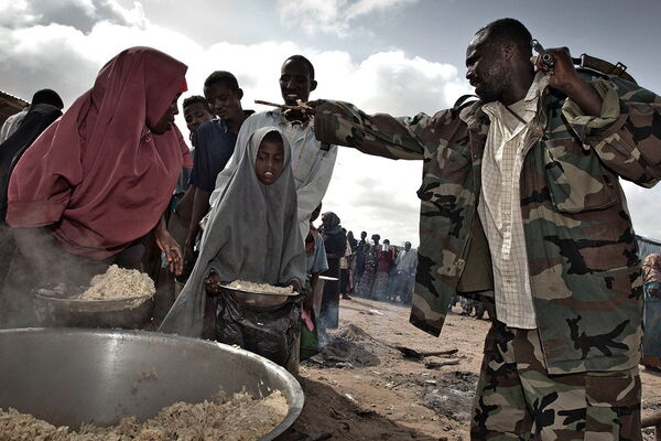A man in camouflage distributes food to people, including women and children, from a large pot outdoors.