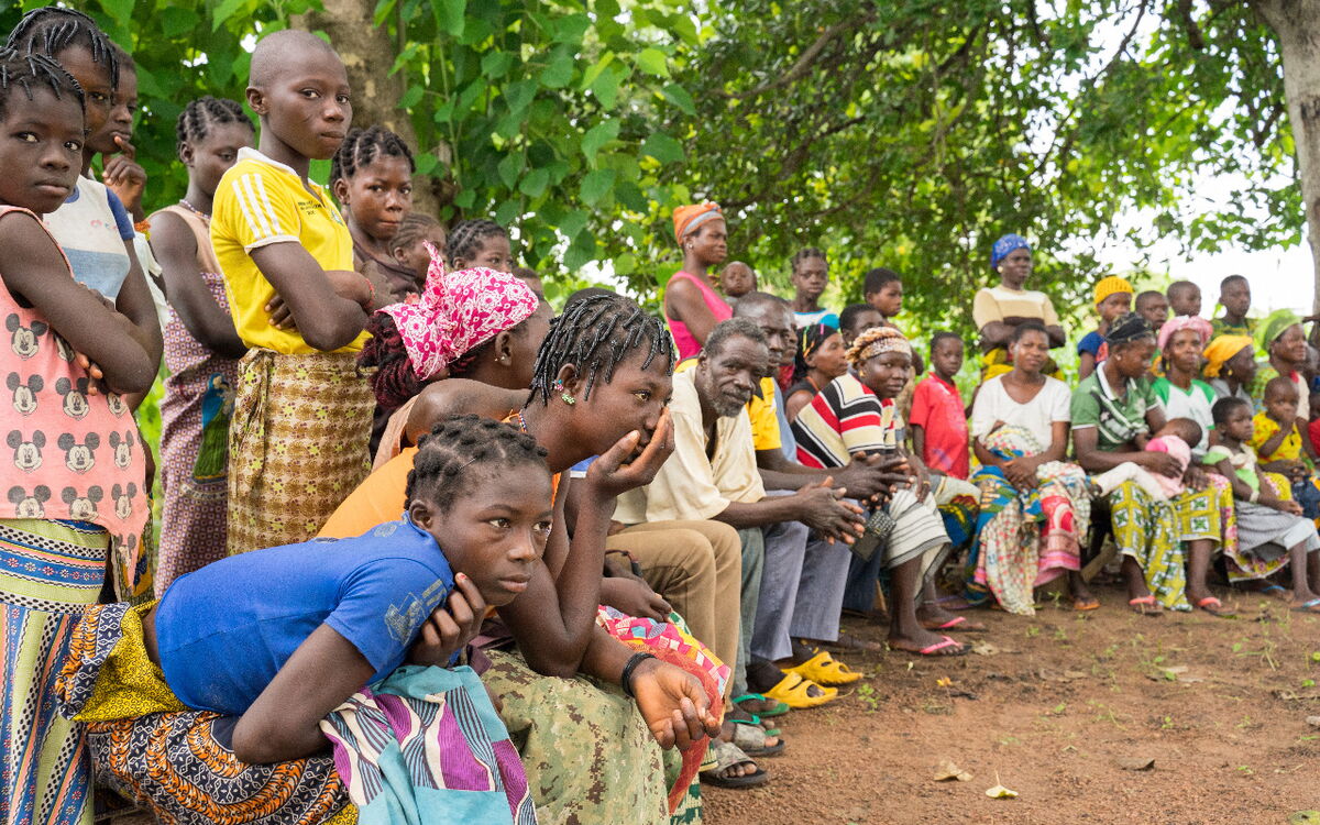 A group of people in colorful clothing gather attentively under trees in Burkina Faso for a community meeting.