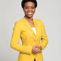 Eine Frau in einem leuchtend gelben Blazer lächelt freundlich in die Kamera vor einem neutralen Hintergrund.