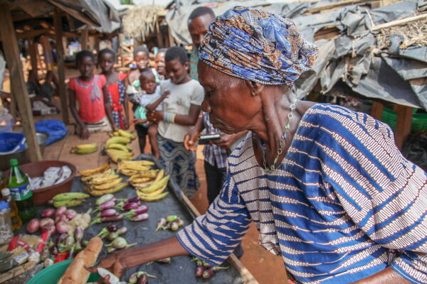An elderly woman in traditional attire selects produce at a bustling outdoor market, surrounded by a group of onlookers.