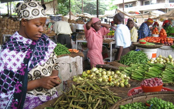 Eine Frau sortiert Gemüse auf einem belebten Marktstand, umgeben von bunten frischen Produkten und anderen Verkäufern.