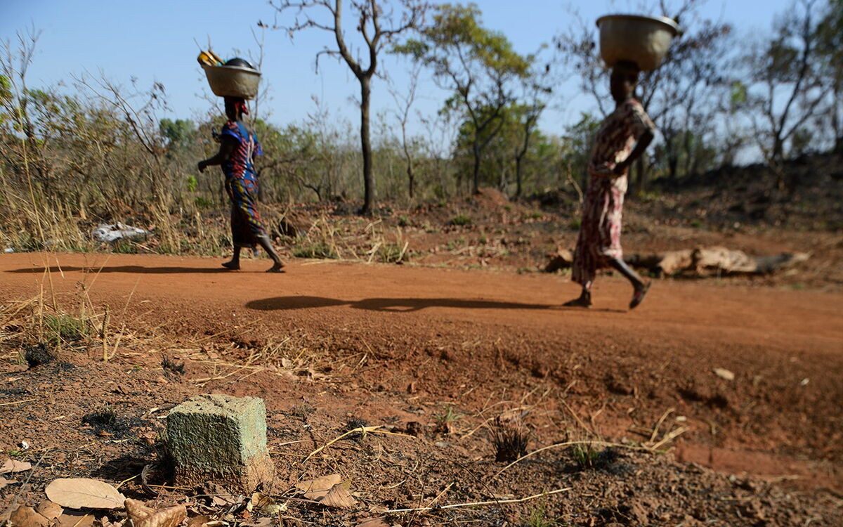 Zwei Frauen tragen Schalen auf dem Kopf entlang eines staubigen Weges in Benin, mit einem Grenzstein im Vordergrund.