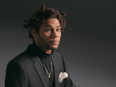 A man with a neat hairstyle and a thoughtful expression wears a black suit and turtleneck against a dark background.