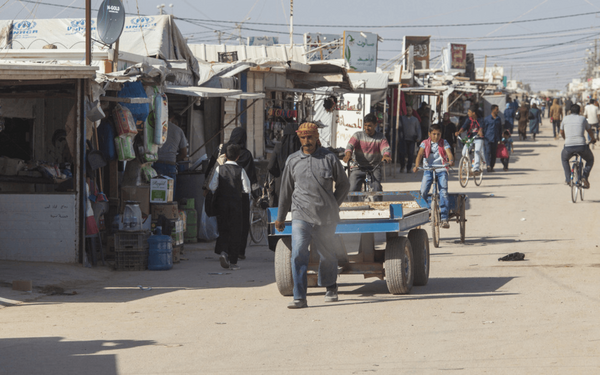 Ein Mann zieht einen Wagen durch einen belebten Markt mit improvisierten Ständen und Fahrradfahrern.