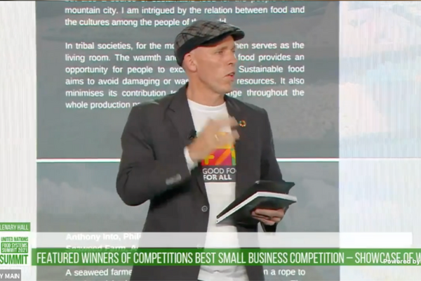A man in a cap and blazer speaks at a podium during a presentation on small business competition winners.