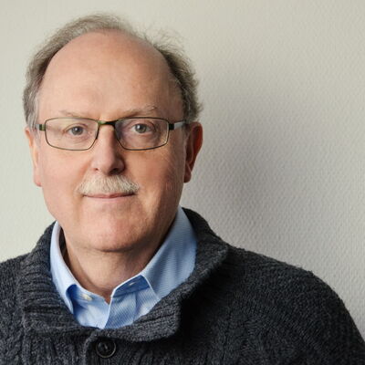 Ein Mann mit Brille und grauem Pullover blickt freundlich in die Kamera vor einem neutralen Hintergrund.