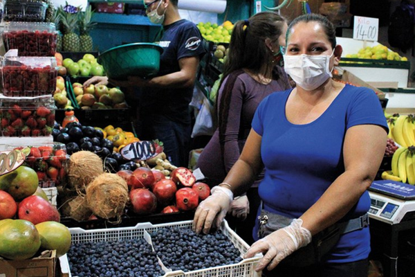 A woman in a blue shirt and mask sells fresh blueberries at a vibrant rural market stand with various fruits.