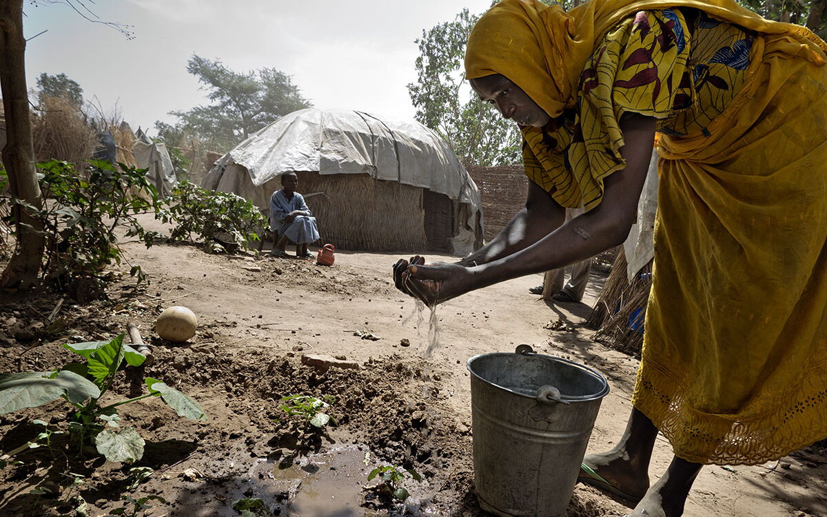 Eine Frau in gelbem Gewand wäscht ihre Hände über einem Eimer, während ein Mann vor einer Hütte sitzt.