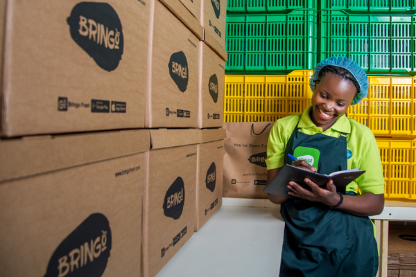 A smiling worker in a green uniform takes notes beside stacked delivery boxes labeled "Bring" in a storage area.