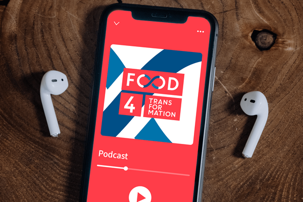 A smartphone displays the "Food 4 Transformation" podcast screen, flanked by wireless earbuds on a wooden surface.