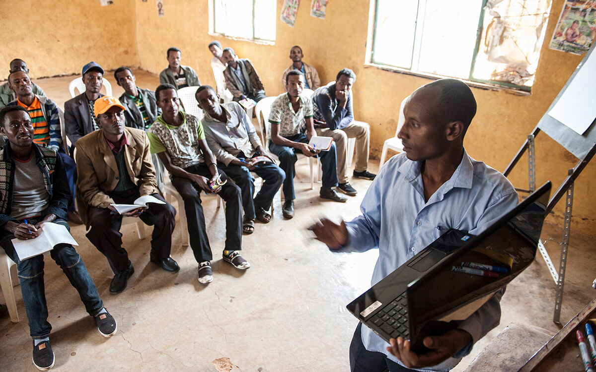 Ein Mann hält einen Vortrag vor einer Gruppe, die aufmerksam in einem Raum mit gelben Wänden zuhört.