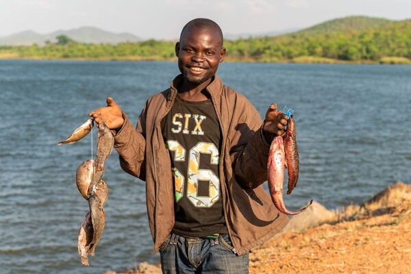 A smiling man stands by a lake, proudly holding freshly caught fish in both hands.