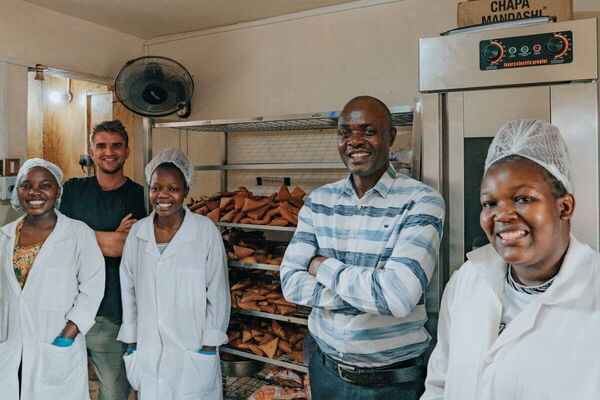A group of five smiling individuals stands in a bakery, showcasing freshly baked goods on metal racks.