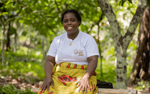 A woman in a white shirt and colorful skirt smiles while sitting outdoors, surrounded by lush greenery, promoting Fairtrade.