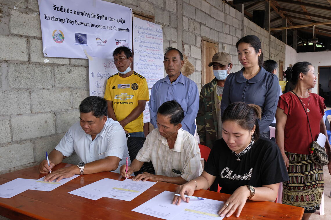 Three villagers sign an agreement with company representatives, observed by others in a community hall setting.