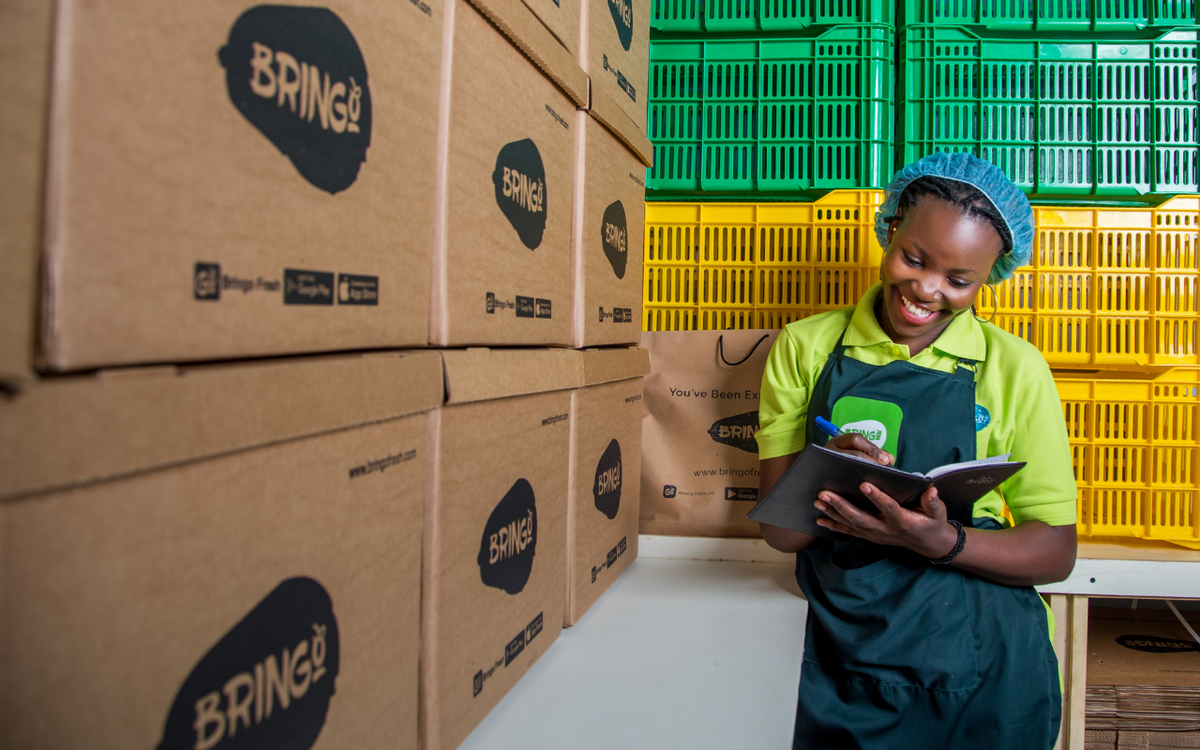 Eine Frau in grüner Arbeitskleidung lächelt und notiert etwas vor gestapelten Kartons und bunten Kisten.