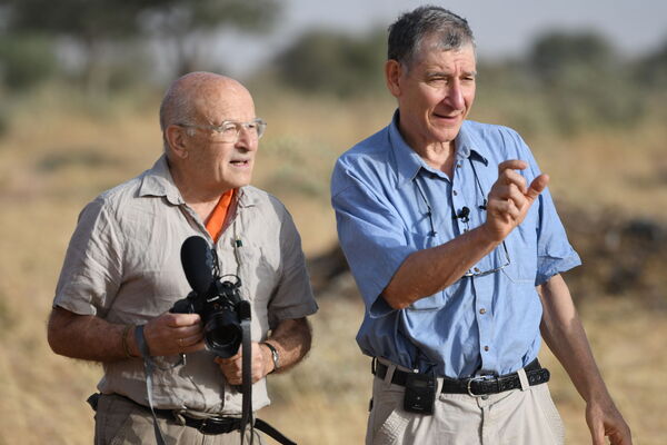 Two men stand in a sunny field in Ghana, one holding a camera and the other gesturing while speaking.