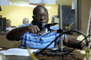 A man in a blue striped shirt speaks passionately into a microphone in a radio studio.