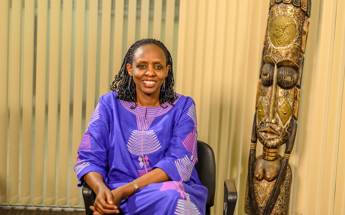 Eine Frau in einem lila Kleid sitzt lächelnd neben einer kunstvoll geschnitzten Holzskulptur.