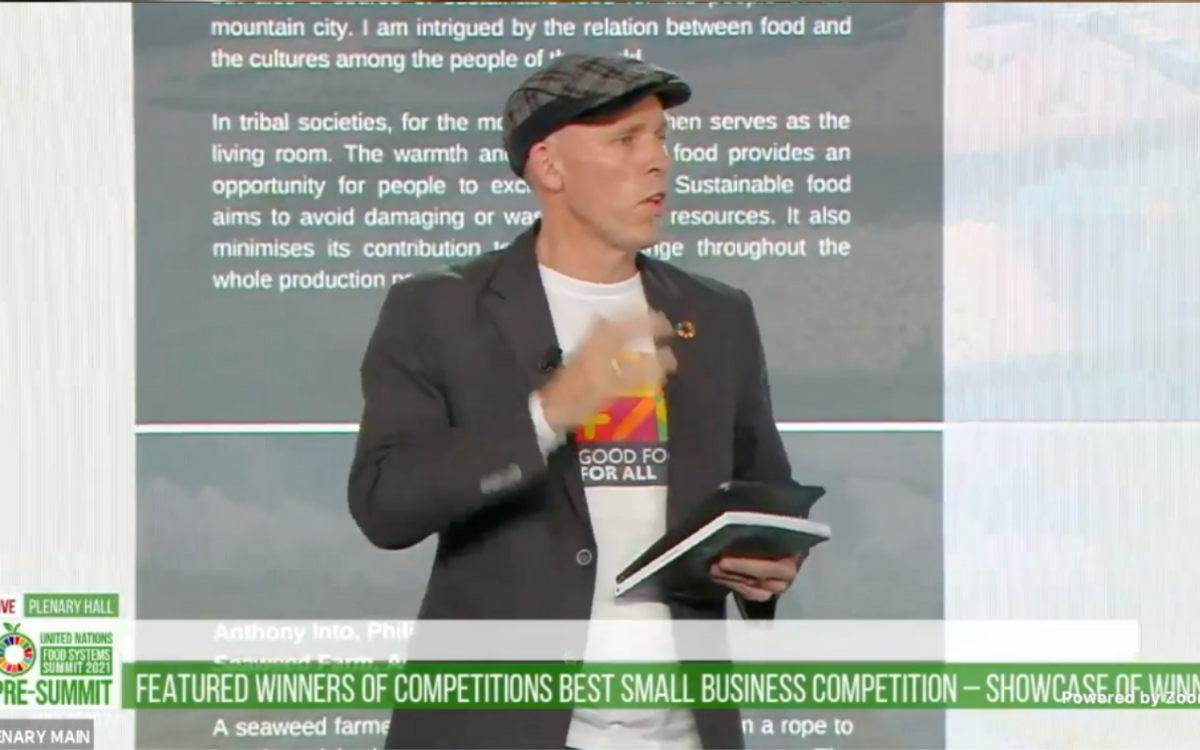 A man in a cap and blazer speaks at a podium during a presentation on small business competition winners.