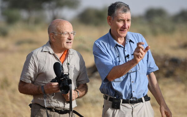 Zwei Männer stehen in der Natur Ghanas, einer hält eine Kamera, während der andere gestikuliert und spricht.