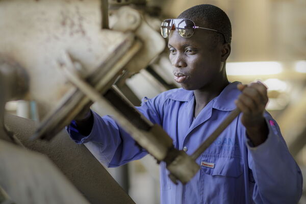 A focused worker in blue coveralls operates machinery, wearing safety goggles on his head.