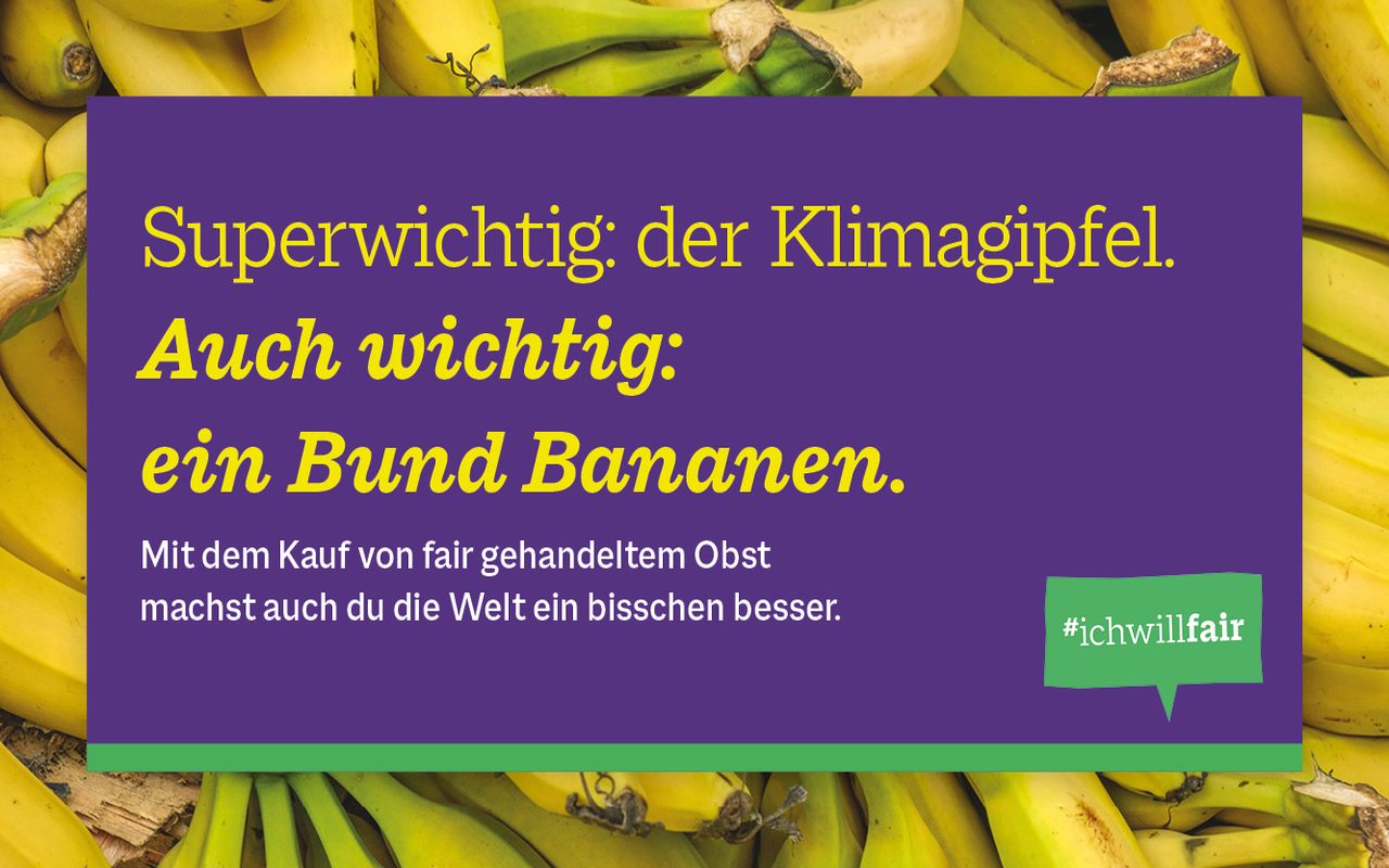 Ein Bündel Bananen liegt hinter einem Schild mit dem Text zur Bedeutung fair gehandelter Produkte und dem Hashtag #ichwillfair.